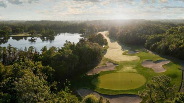 Aerial view of a lush golf course surrounded by trees and water under a bright morning sky.