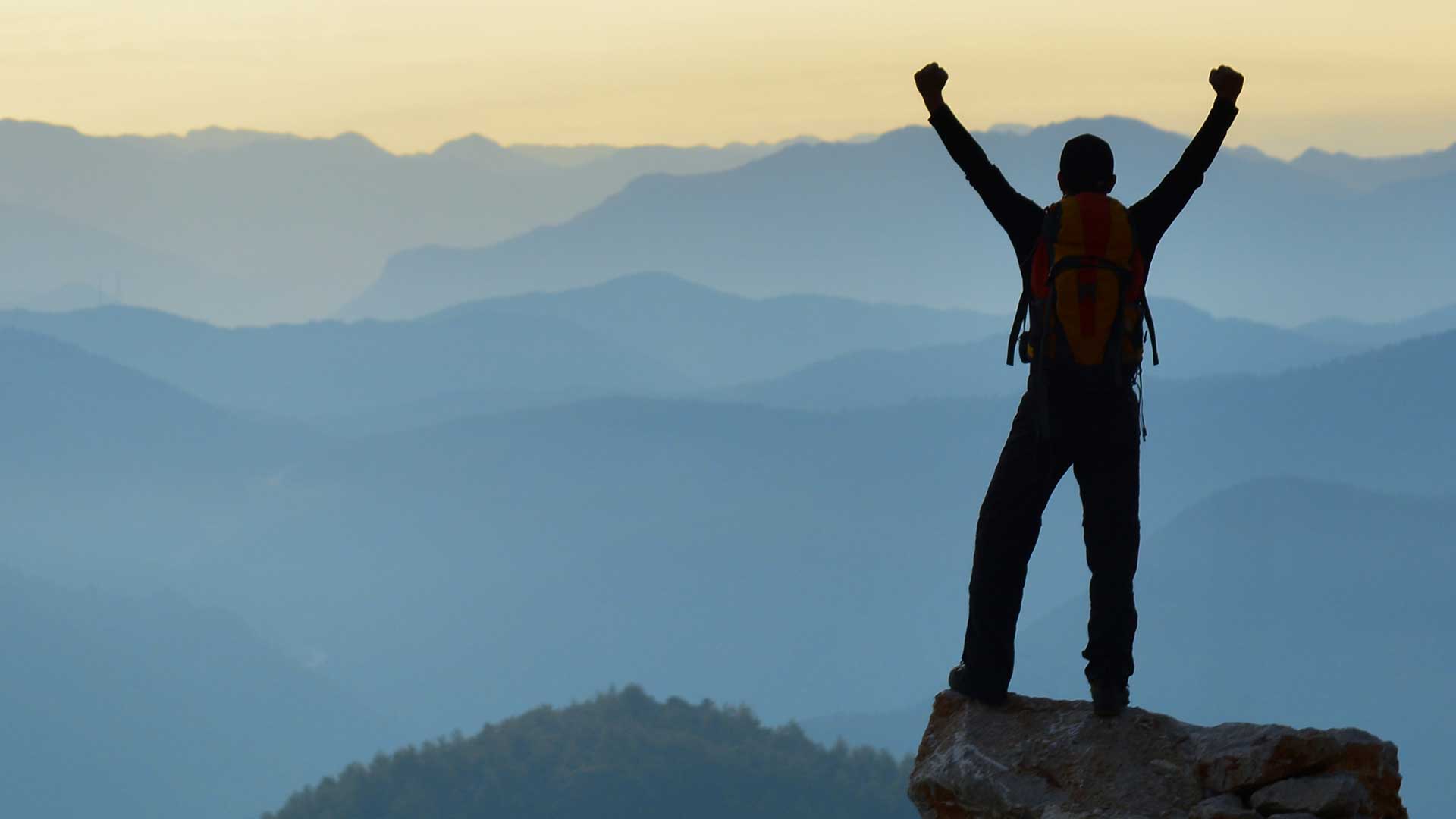 Person standing triumphantly on a mountain peak at sunrise, overlooking layers of misty blue mountains.