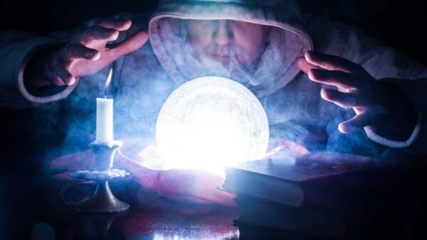 A hooded figure gazes into a glowing crystal ball surrounded by smoke, with a lit candle and books nearby.