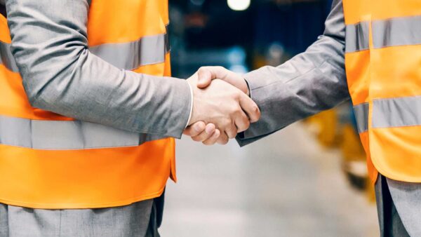 Two workers wearing orange safety vests shaking hands inside an industrial facility.