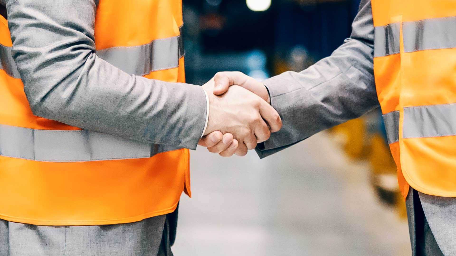Two workers wearing orange safety vests shaking hands inside an industrial facility.