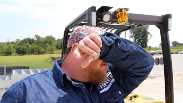 Worker wiping sweat from his face outdoors, showing signs of heat stress while operating equipment.