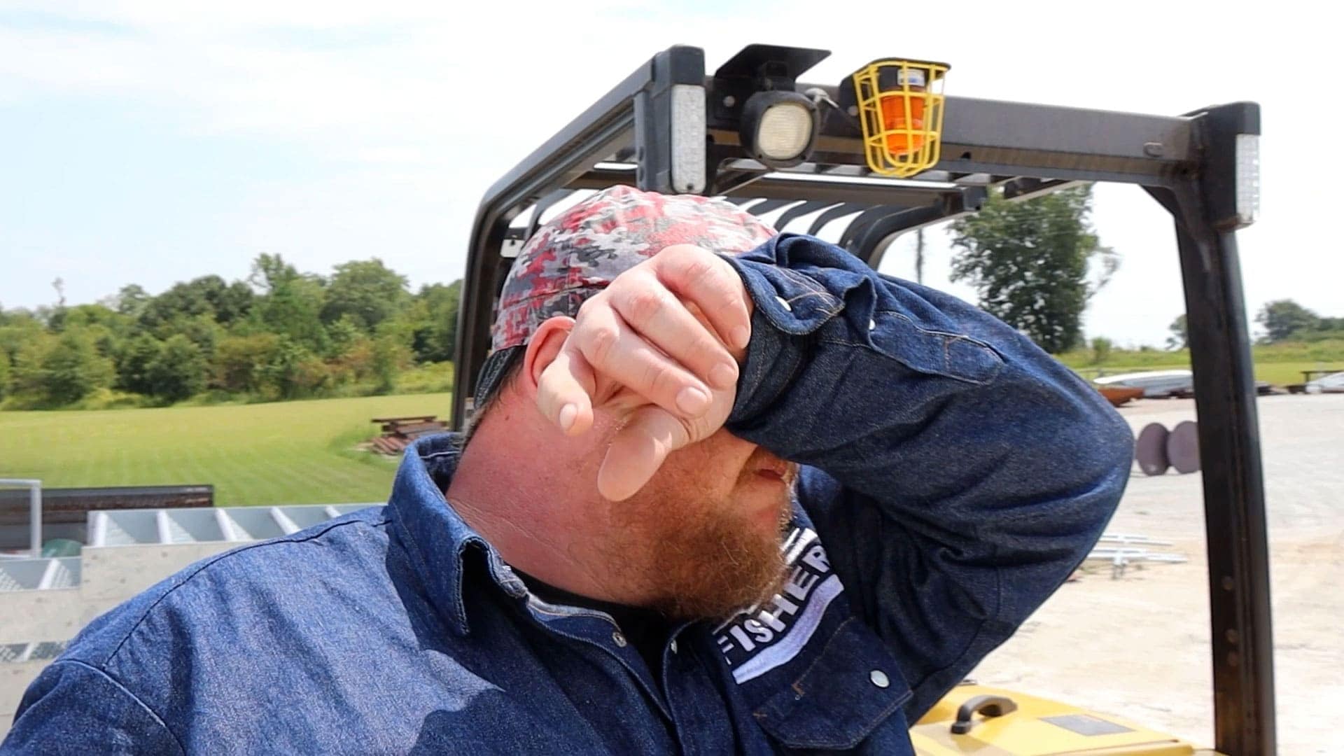 Worker wiping sweat from his face outdoors, showing signs of heat stress while operating equipment.