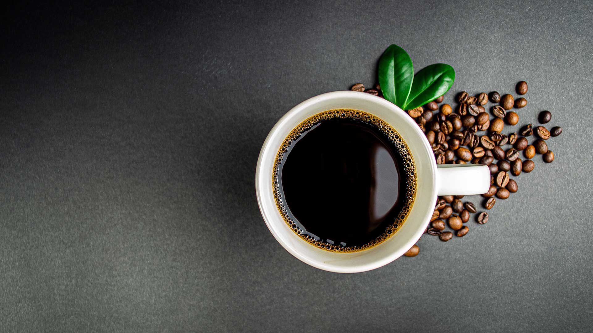 Cup of black coffee surrounded by roasted coffee beans on a dark surface.
