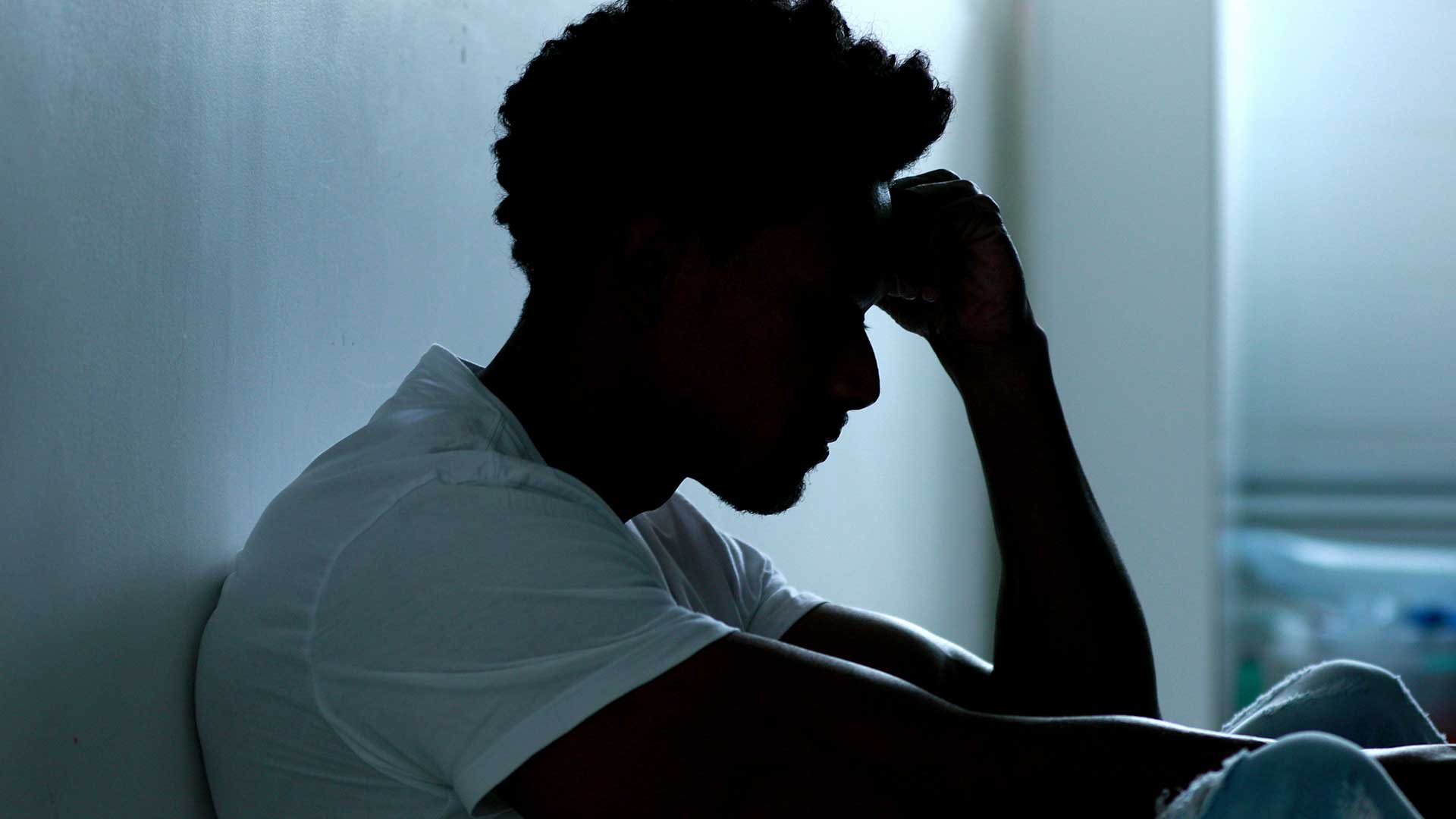 Silhouette of a person sitting against a wall, head resting on their hand, appearing distressed or deep in thought.