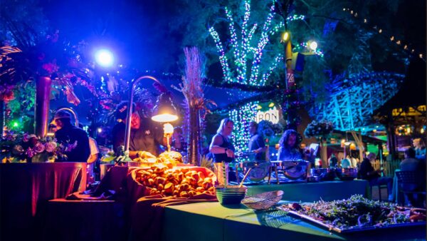 Outdoor evening buffet with guests dining under colorful lights and illuminated trees at a theme park setting.