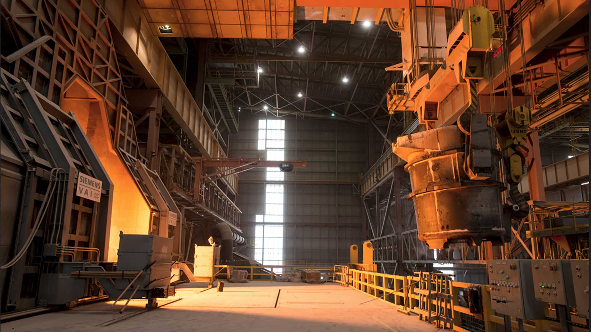 Interior view of a stainless steel production facility with heavy industrial equipment, overhead cranes, and steelmaking machinery.